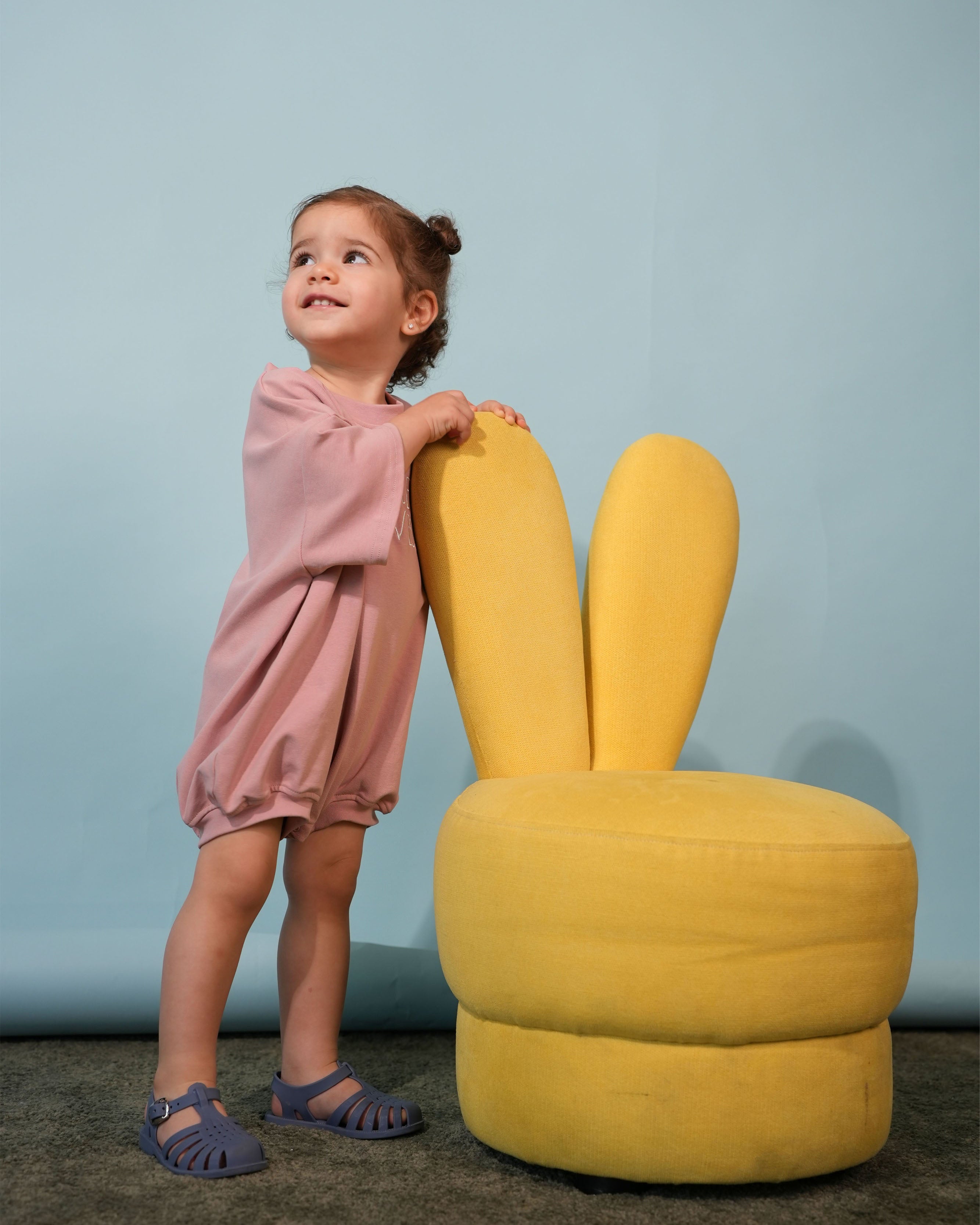 lifestyle shot of a baby wearing the embroidered blush pink romper in a sunlit room standing next to a yellow bunny chair.