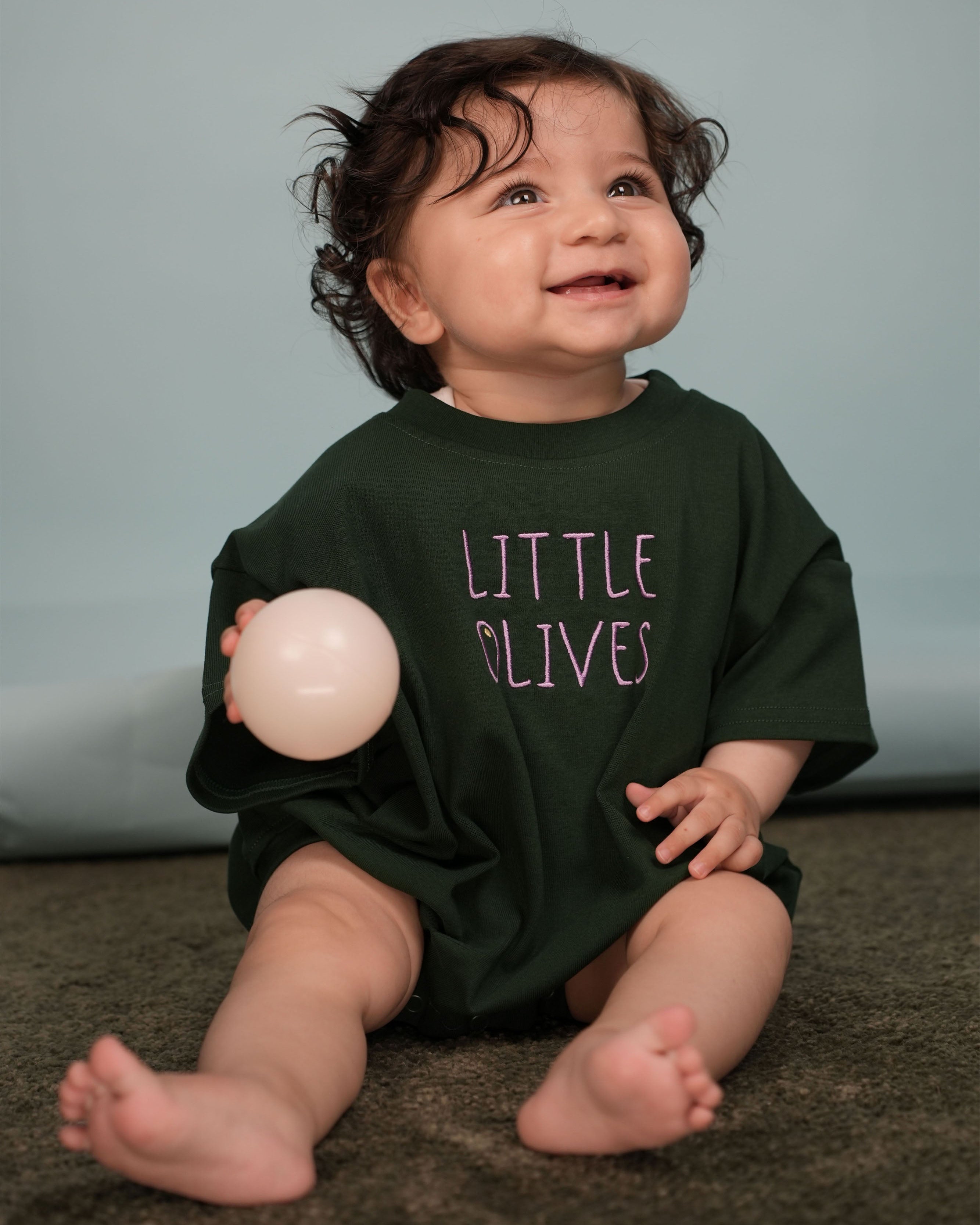 Lifestyle shot of a baby looking like "tiny royalty" in the green and purple embroidered romper.