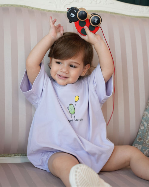 Smiling child wearing a lilac romper with a cartoon design, sitting on a couch holding a toy.